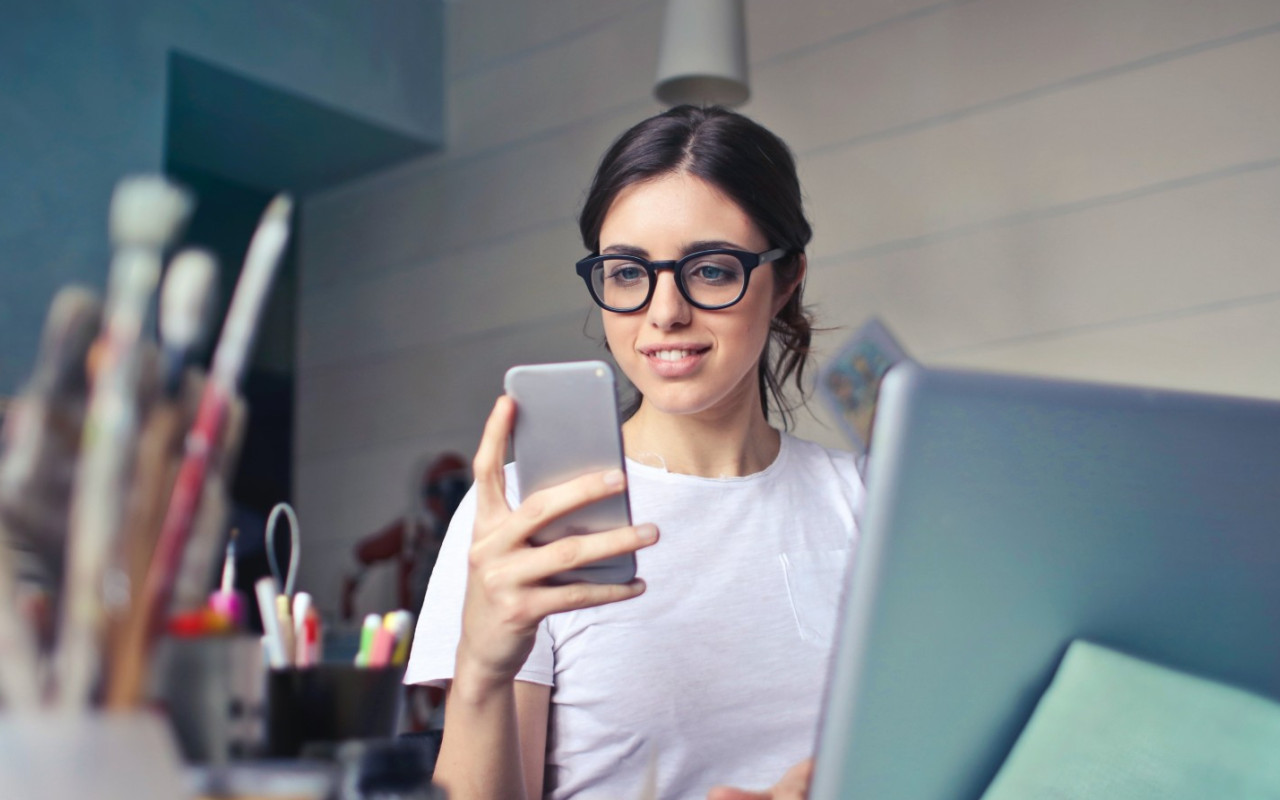 Young woman smiling at her phone while sitting at a desk surrounded by artistic tools