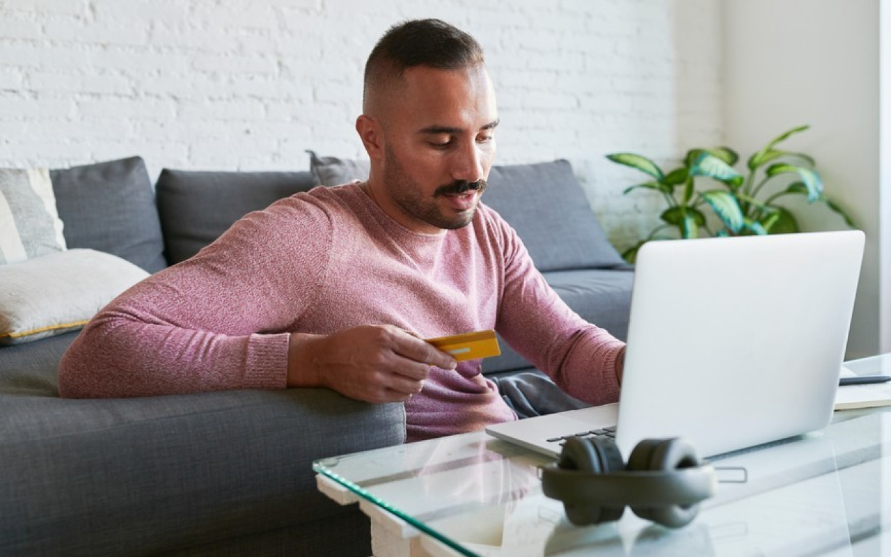 Young man sitting on floor in front of laptop holding credit card, in living room.